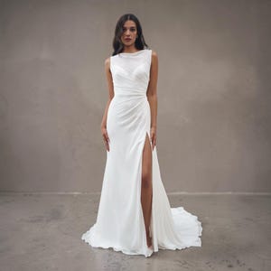 Puede incluir: Vestido de novia blanco sin mangas con cuello alto y abertura en el muslo. El vestido presenta un corpiño drapeado y una falda fluida que se arrastra por el suelo. El logotipo "Versalles" es visible.