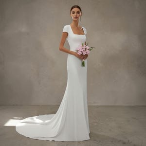 May include: A white wedding dress with a square neckline and short sleeves. The dress has a fitted bodice and a flowing skirt that extends into a long train. The person is holding a bouquet of pink flowers. The logo "Versailles" is in the upper left corner.