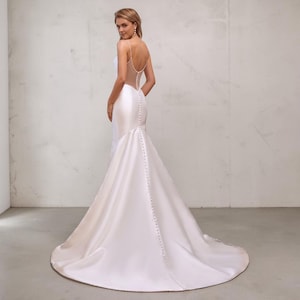 Pode incluir: Um vestido longo de cetim branco com costas transparentes, alças finas e um corpete justo que se transforma numa saia rodada com cauda. Uma fila vertical de pequenos botões brancos percorre o centro da parte de trás do vestido.