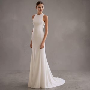 May include: A white, floor-length wedding gown with a high neckline and a fitted silhouette. The dress features a slight train and a simple, elegant design. The model is standing in front of a gray background.