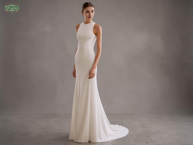 May include: A white, floor-length wedding gown with a high neckline and a fitted silhouette. The dress features a slight train and a simple, elegant design. The model is standing in front of a gray background.