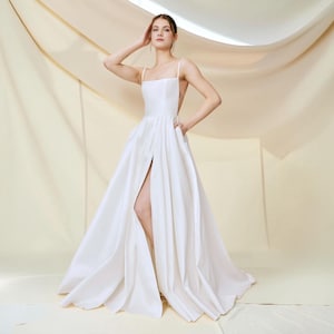 May include: A floor-length white gown featuring a square neckline, thin spaghetti straps, and a full skirt with a high front slit. The dress has a fitted bodice and a flowing, pleated skirt design. The logo Versailles is visible in the top left corner.
