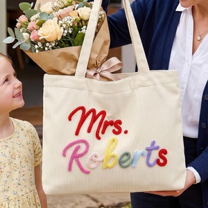 Puede incluir: Un bolso tote de pana color crema con el texto bordado Mrs. Roberts en letras de hilo coloridas y en relieve. El bolso tiene dos correas para el hombro y una forma rectangular, con el nombre mostrado en rojo, rosa, amarillo, verde y azul.