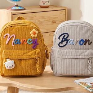 May include: A mustard yellow corduroy backpack with the name Nancy embroidered in rainbow colors and a small flower patch, and a light gray corduroy backpack with the name Baron embroidered in dark blue, both featuring front pockets and braided zipper pulls.