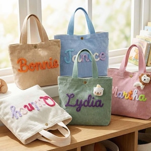 May include: A collection of corduroy tote bags in various colors, each embroidered with a name. The bags have matching handles and are displayed on a wooden shelf near a window. A wooden toy car is also visible.
