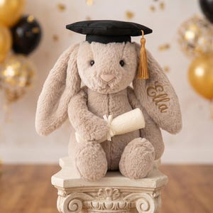 May include: A plush bunny wearing a black graduation cap with a gold tassel, holding a rolled-up scroll. The bunny is light brown and has the name "Ella" embroidered on its ear. It is sitting on a decorative white pedestal, with balloons in the background.