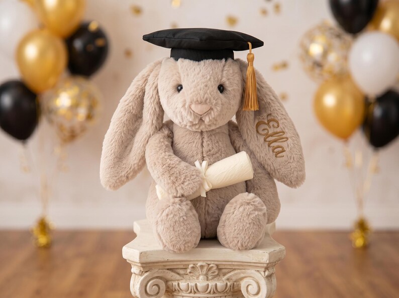 May include: A plush bunny wearing a black graduation cap with a gold tassel, holding a rolled-up scroll. The bunny is light brown and has the name "Ella" embroidered on its ear. It is sitting on a decorative white pedestal, with balloons in the background.