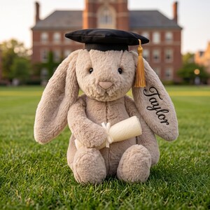 May include: A plush bunny wearing a black graduation cap with a gold tassel and holding a rolled-up diploma tied with a ribbon. The bunny's ear is embroidered with the name "Taylor". The bunny is sitting on green grass.