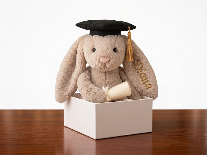 May include: A plush bunny wearing a black graduation cap with a gold tassel, holding a rolled-up scroll. The bunny is inside a white box, with the name "Diana" embroidered on its ear. The bunny is a light brown color.