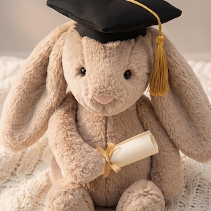 May include: A plush, light brown bunny wearing a black graduation cap with a gold tassel. The bunny holds a rolled-up diploma tied with a gold ribbon. The soft toy is sitting on a white, textured blanket.