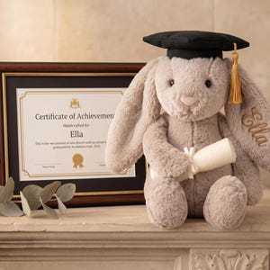 May include: A plush, gray bunny wearing a black graduation cap with a gold tassel, holding a rolled diploma. The bunny has "Ella" embroidered on its ear. A framed certificate of achievement is in the background.