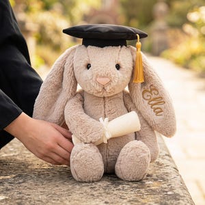 May include: A plush, beige bunny wearing a black graduation cap with a gold tassel. The bunny holds a rolled-up diploma and has the name "Ella" embroidered on its ear. The bunny is sitting on a stone surface.