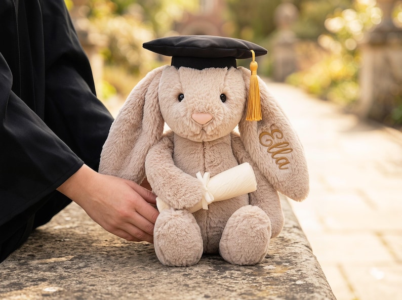 May include: A plush, beige bunny wearing a black graduation cap with a gold tassel. The bunny holds a rolled-up diploma and has the name "Ella" embroidered on its ear. The bunny is sitting on a stone surface.