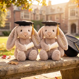 May include: Two plush bunny rabbits wearing graduation caps and holding diplomas. The bunnies are light brown with long ears and are sitting on a stone bench. One bunny has "Sarah" embroidered on its ear, the other has "Jessica". A graduation cap with a gold tassel is next to them.