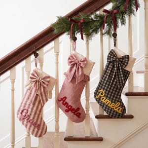May include: Three decorative Christmas stockings hanging from a staircase railing. The first is red and white striped with the name Grace, the second is solid dusty pink with the name Angela, and the third is black and tan plaid with the name Pamela.