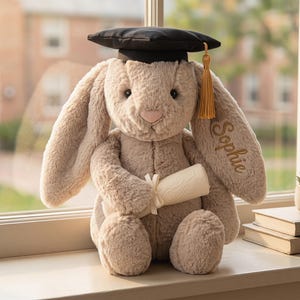 May include: A plush, beige graduation bunny wearing a black mortarboard with a gold tassel. The bunny holds a rolled-up diploma and has the name "Sophie" embroidered on its ear. The bunny is sitting on a windowsill.