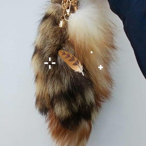 Può includere: Un portachiavi a forma di coda in pelliccia sintetica soffice con un mix di fibre marroni, nere, color cuoio e bianche. La coda è fissata a un moschettone e una catena in metallo dorato, decorata con una piccola icona grafica di piuma marrone.