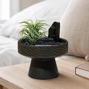 May include: A dark gray pedestal bowl with a textured exterior containing a green air plant and several jagged black tourmaline crystals. The bowl has a wide, shallow basin and a sturdy, tapered base, filled with small black decorative stones.