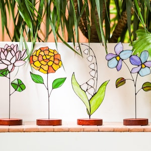 Op de afbeelding: Vier kleurrijke glas-in-lood bloemensculpturen. De bloemen zijn een roze lotus, een geel-oranje goudsbloem, een witte lelietje-van-dalen en blauwe en paarse viooltjes. Elke bloem is gemonteerd op een dunne zwarte metalen steel en een kleine, ronde houten basis.