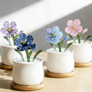 Op de afbeelding: Vier glazen bloemensculpturen in witte keramische potten met houten voetjes. De bloemen zijn in tinten blauw, roze en paars, met gele centra en groene stelen en bladeren. De potten zijn gevuld met witte kiezelstenen.