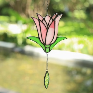 May include: A stained glass suncatcher shaped like a pink tulip flower with green leaves, held together by dark metal solder lines. A small, textured green glass leaf-shaped pendant hangs from the bottom of the flower on a thin metal chain.