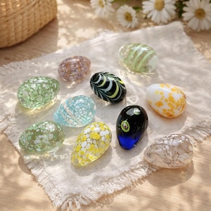 May include: A collection of decorative glass egg-shaped ornaments in various colors and patterns. The eggs feature speckled, striped, and swirled designs in shades of green, blue, yellow, and brown. They rest on a white linen cloth.