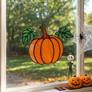 May include: A stained glass pumpkin decoration with green leaves hangs in front of a window. The orange pumpkin is outlined in black and has a stem. A spider and a web are visible in the window.