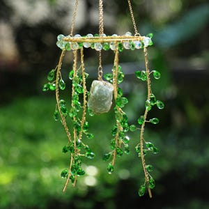 May include: A hanging decorative piece featuring a gold-colored metal frame adorned with green crystal beads and a central, light green stone. Delicate strands of green beads cascade downwards, creating a natural, organic aesthetic. The piece is suspended by a gold chain.