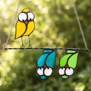 Op de afbeelding: Een glas-in-lood suncatcher met drie vogels op een draad. Eén gele vogel zit rechtop op de draad, terwijl een blauwgroene en een limoengroene vogel ondersteboven aan de draad hangen. De vogels hebben grote witte ogen en zwarte metalen randen.