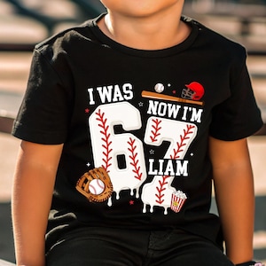 May include: Black t-shirt with a baseball-themed design. The shirt features the text "I WAS 6, NOW I'M 7 LIAM" with baseball graphics, including a bat, ball, helmet, and glove. The number 6 and 7 are designed to look like baseballs.