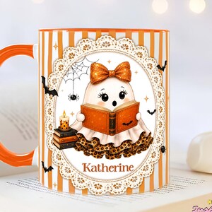 May include: A white ceramic mug with an orange striped pattern and an orange handle. The center features a circular lace frame containing a ghost reading an orange book, wearing a glittery orange bow and leopard print skirt, with the name Katherine.