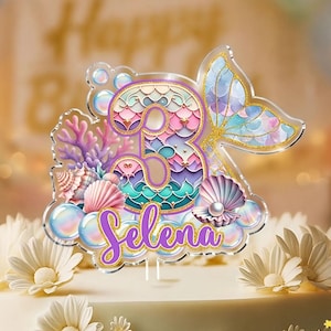 Puede incluir: Un adorno para tarta con tem&aacute;tica de sirena. Presenta el n&uacute;mero 3, una cola de sirena, conchas marinas y el nombre "Selena" en escritura p&uacute;rpura. El dise&ntilde;o incorpora colores pastel y detalles dorados, ideal para una celebraci&oacute;n de cumplea&ntilde;os.
