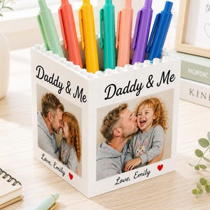 May include: A white pen holder with a photo of a father and child, the words "Daddy & Me" and "Love, Emily" are printed on the holder. The holder contains several colorful pens in shades of green, orange, yellow, teal, pink, purple, and blue.