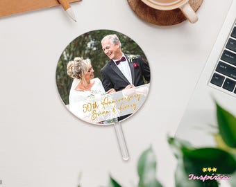 May include: A circular acrylic cake topper featuring a printed photograph of a couple in formal wedding attire. The text 50th Anniversary Brian & Avery is written in gold cursive script across the bottom portion of the circular topper.