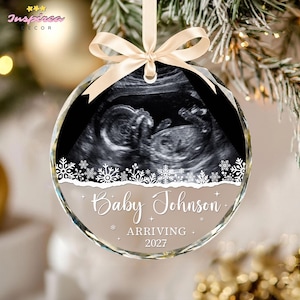 May include: A round, clear glass Christmas ornament featuring a black and white ultrasound image of a fetus. The ornament is decorated with white snowflakes and the text Baby Johnson ARRIVING 2027, finished with a cream-colored ribbon bow.