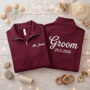 May include: Two burgundy quarter-zip sweatshirts. The left sweatshirt features the embroidered text "Mr. Jones" on the chest. The right sweatshirt displays the embroidered text "Groom" above the date "29.5.2026" on the back.