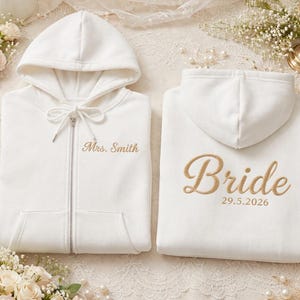 Könnte beinhalten: Zwei weiße Kapuzenpullover mit Reißverschluss und goldener Stickerei. Der linke Pullover hat eine Kapuze, Kordelzüge und den Text Mrs. Smith auf der Brust. Der rechte Pullover hat eine Kapuze und den Text Bride mit dem Datum 29.5.2026 auf dem Rücken.