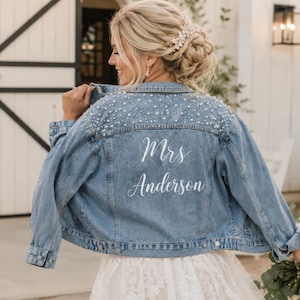 May include: A light blue denim jacket featuring white pearl embellishments across the upper back and shoulders. The back of the jacket displays the white text Mrs. Anderson in a cursive script font. The jacket has long sleeves and a collar.
