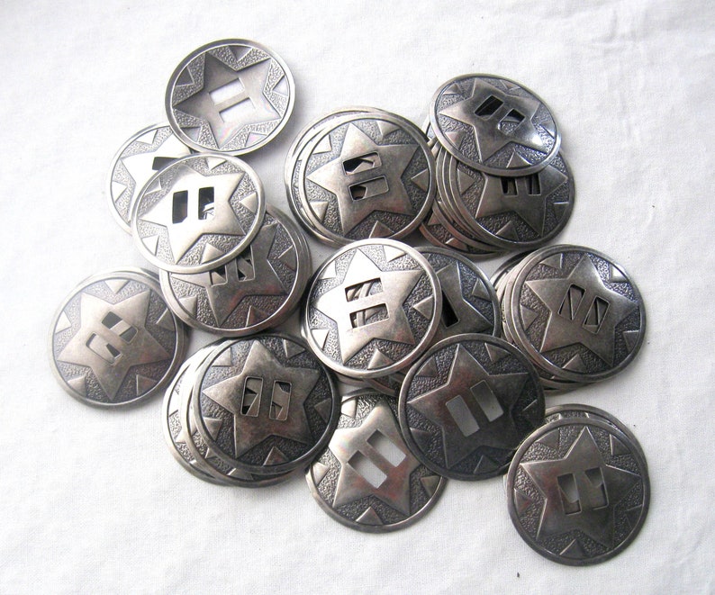 May include: A pile of silver-toned metal conchos with a star design and two slits. The conchos are approximately 1 inch in diameter.