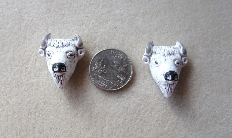 May include: Two white ceramic buffalo head figurines with black noses and gray horns. The figurines are about the size of a US quarter.