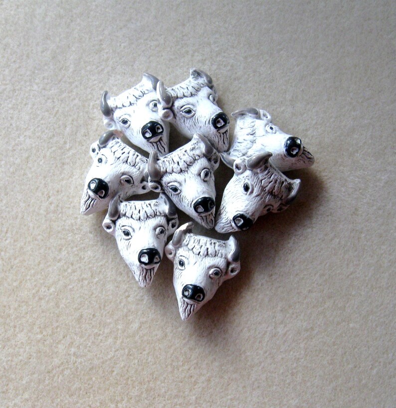 May include: A group of nine white ceramic buffalo head charms with black painted details. The charms are arranged in a cluster on a light beige surface.