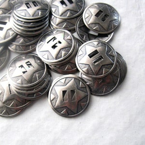 May include: A pile of silver-toned metal star-shaped buttons with a double-line design in the center. The buttons are stacked and scattered on a white surface.