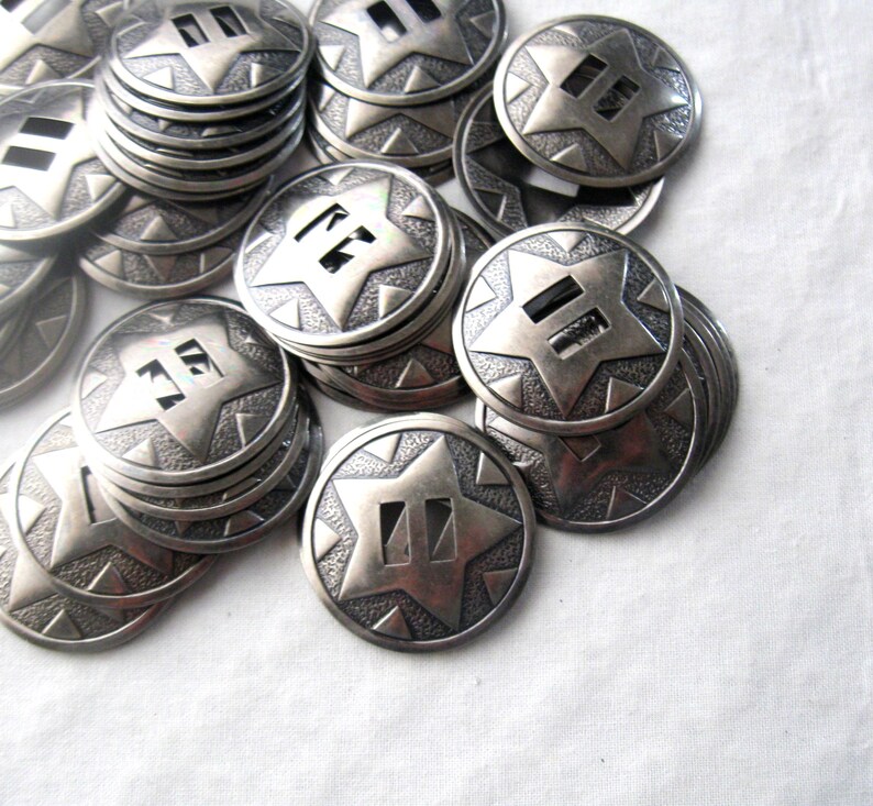 May include: A pile of silver-toned metal star-shaped buttons with a double-line design in the center. The buttons are stacked and scattered on a white surface.