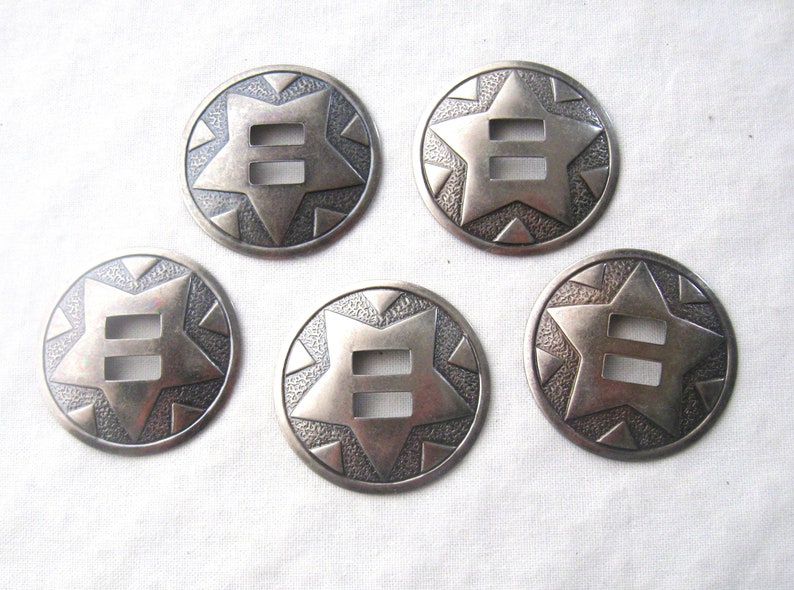 May include: Six silver-toned metal conchos with a star design and a double-slot opening. The conchos have a textured surface and are perfect for adding a western touch to your projects.
