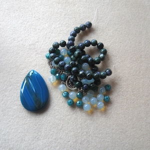 May include: A collection of jewelry components, including a large, teardrop-shaped blue agate pendant with striped patterns. There are also strands of blue, green, and white beads, along with a silver-toned metal component.