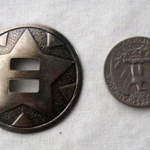 May include: A silver-toned metal concho with a star design and two cut-out slots. The concho is about the size of a US quarter.
