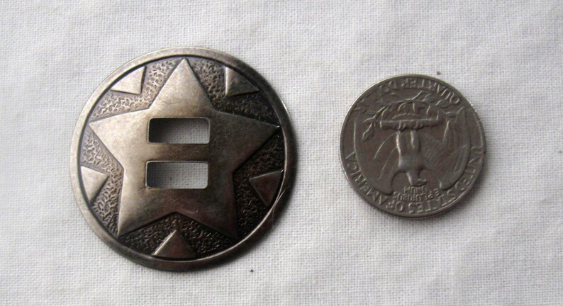 May include: A silver-toned metal concho with a star design and two cut-out slots. The concho is about the size of a US quarter.