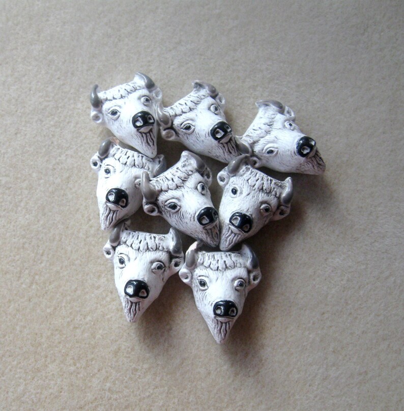 May include: Eight white ceramic buffalo head beads with black eyes and nostrils. The beads are arranged in a heart shape.