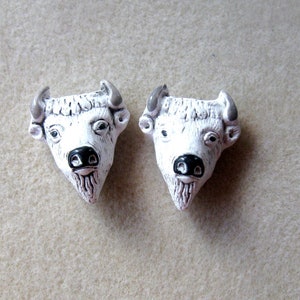 May include: Two white ceramic buffalo head sculptures with black noses and gray horns. The sculptures are facing forward and are on a light brown surface.