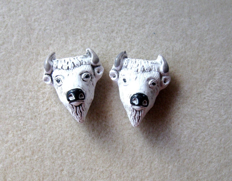 May include: Two white ceramic buffalo head sculptures with black noses and gray horns. The sculptures are facing forward and are on a light brown surface.